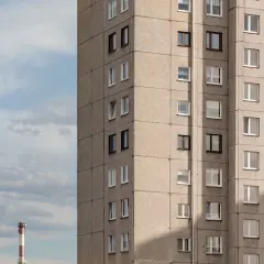 Szary blok mieszkalny z wieloma oknami, przypominający budynek z mieszkaniami socjalnymi. W tle błękitne niebo z chmurami i czerwono-biały komin.