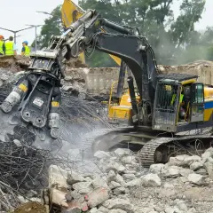 Maszyna budowlana wykonuje rozbiórkę, krusząc beton i stal. W tle widać pracowników w kamizelkach.