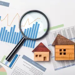 Analiza wykresów i tabeli z cenami materiałów budowlanych, z lupą i modelami domów.