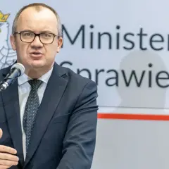 Mężczyzna w garniturze przemawia przy mikrofonie na tle godła Polski i napisu "Ministerstwo Sprawiedliwości". Może mówić o problemach z mieszkaniem komunalnym w Warszawie.