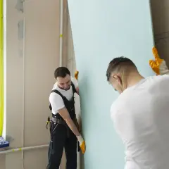 Dwóch budowlańców montuje płyty regipsowe. Jeden z nich przytrzymuje duży, jasnoniebieski arkusz, podczas gdy drugi ustawia go w odpowiednim miejscu.