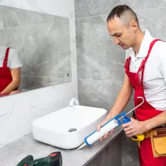 Fachowiec w czerwonym ogrodniczkach nakłada silikon wokół umywalki. Czas, ile schnie silikon, zależy od warunków.