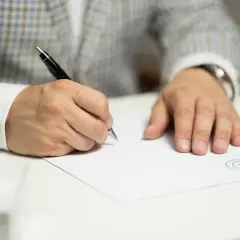 Mężczyzna w eleganckim garniturze podpisuje umowę deweloperską. Na dokumencie widnieje logo.