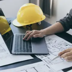 Architekt analizuje plany, obliczając powierzchnię użytkową budynku na laptopie. Obok leży żółty kask.