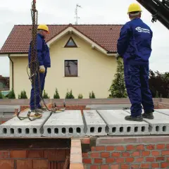 Dwóch robotników w żółtych kaskach i niebieskich kombinezonach montuje strop filigran na budowie domu.