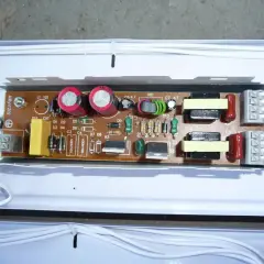 Elektroniczny statecznik z kondensatorami, cewkami i innymi elementami na płytce drukowanej, podłączony białymi przewodami.