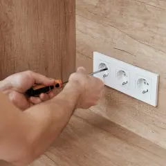 Ręce majsterkowicza dokręcają śrubki w białej ramce z trzema gniazdkami, zapewniając bezpieczne napięcie w gniazdku.