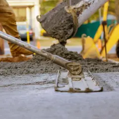 Pracownik wylewa beton na chudziaka, używając łaty do wyrównania powierzchni. W tle widać koparkę.