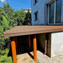 Nowy taras na słupach betonowych, z brązową, drewnopodobną nawierzchnią, otoczony zielenią.