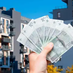 Dłoń trzyma polskie banknoty 100 zł na tle nowoczesnych bloków mieszkalnych. Szybka sprzedaż mieszkania przed upływem 5 lat.