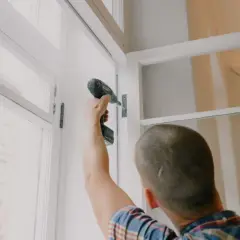 Mężczyzna przykręca zawias w nowym oknie. Planujesz wymianę okien w bloku? Cena może być niższa niż myślisz!