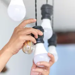 Ręce wymieniają żarówkę w lampie wiszącej. Widać starą żarówkę i nową LED.