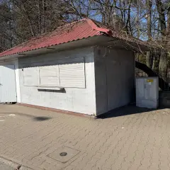 Mały lokal użytkowy z czerwoną, falistą dachem i białymi okiennicami. Obok stoi hulajnoga elektryczna.