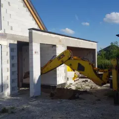 Żółta koparko-ładowarka pracuje przy budowie domu z bloczków. Widać okna i drzwi. Trwa wlz.