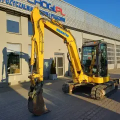 Żółta koparka Komatsu przed budynkiem. Zastanawiasz się, ile kosztuje godzina pracy koparki? Sprawdź ofertę wypożyczalni!
