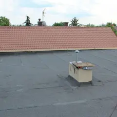 Płaski dach z papy, na którym stoi ceglany ogniomur z metalową rurą. W tle dach skośny pokryty dachówką.