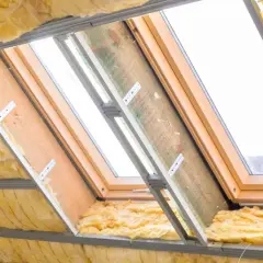 Ocieplenie poddasza wełną mineralną w trakcie prac. Widoczne profile metalowe, drewniana konstrukcja okna dachowego i żółta wełna.