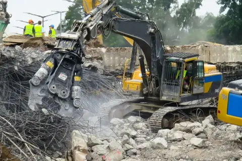 Maszyna budowlana wykonuje rozbiórkę, krusząc beton i stal. W tle widać pracowników w kamizelkach.