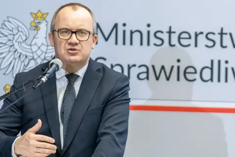 Mężczyzna w garniturze przemawia przy mikrofonie na tle godła Polski i napisu "Ministerstwo Sprawiedliwości". Może mówić o problemach z mieszkaniem komunalnym w Warszawie.