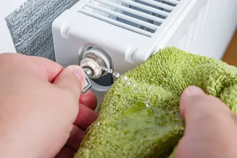 Ręka z kluczykiem odpowietrza grzejnik zimny na dole. Woda kapie na zielony ręcznik.