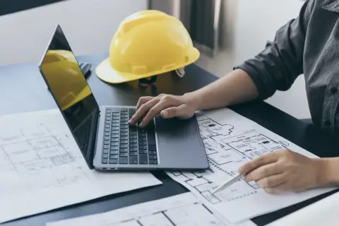 Architekt analizuje plany, obliczając powierzchnię użytkową budynku na laptopie. Obok leży żółty kask.