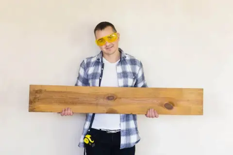 Mężczyzna w okularach ochronnych trzyma drewnianą deskę, która może posłużyć jako panele podłogowe na ścianie.