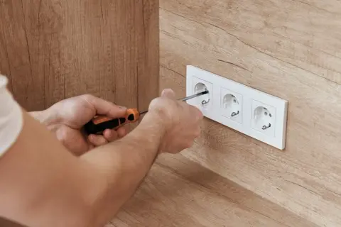 Ręce majsterkowicza dokręcają śrubki w białej ramce z trzema gniazdkami, zapewniając bezpieczne napięcie w gniazdku.