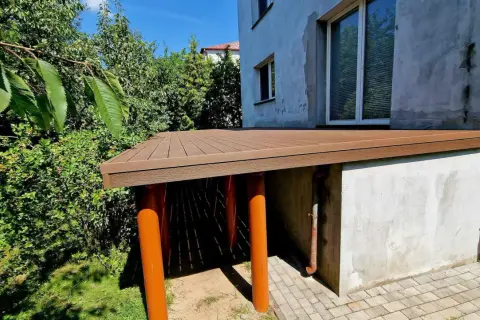 Nowy taras na słupach betonowych, z brązową, drewnopodobną nawierzchnią, otoczony zielenią.