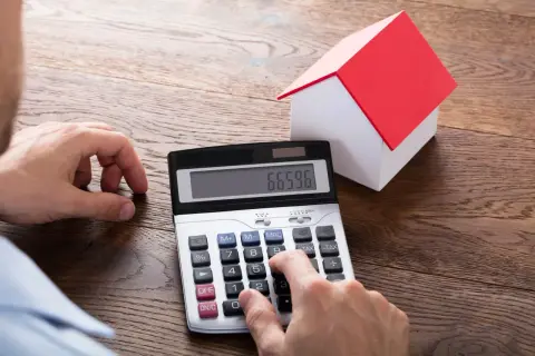 Ręce liczą na kalkulatorze, obok stoi model domu. Obliczenia mogą dotyczyć podatku od działki budowlanej.