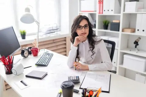 Kobieta w okularach siedzi przy biurku z dokumentami, kalkulatorem i kubkiem kawy.