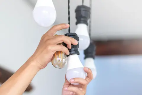 Ręce wymieniają żarówkę w lampie wiszącej. Widać starą żarówkę i nową LED.