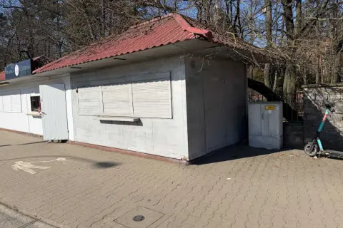 Mały lokal użytkowy z czerwoną, falistą dachem i białymi okiennicami. Obok stoi hulajnoga elektryczna.