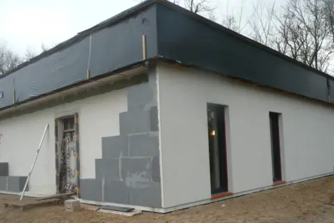 Budowa domu ze styropianu w toku. Ściany z bloczków, częściowo otynkowane, dach przykryty czarną folią.