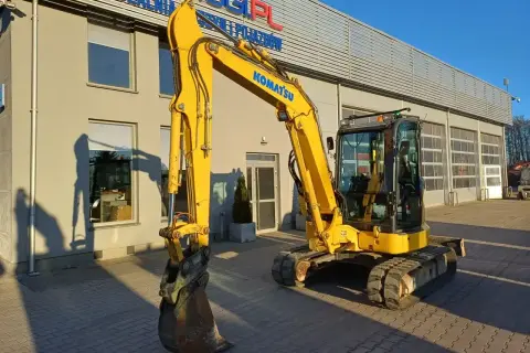 Żółta koparka Komatsu przed budynkiem. Zastanawiasz się, ile kosztuje godzina pracy koparki? Sprawdź ofertę wypożyczalni!