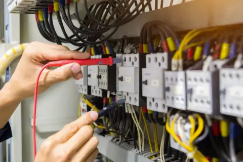 Elektryk sprawdza przewody w szafie sterowniczej. Widać multimetr i liczne kable, w tym przewód neutralny.