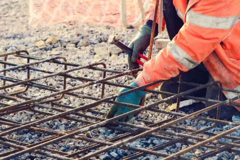 Budowlaniec w pomarańczowej kamizelce z odblaskami pracuje nad zbrojeniem rozproszonym, łącząc stalowe pręty.