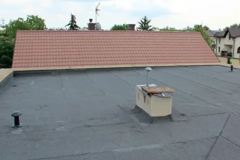 Płaski dach z papy, na którym stoi ceglany ogniomur z metalową rurą. W tle dach skośny pokryty dachówką.