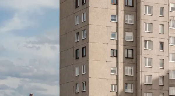 Szary blok mieszkalny z wieloma oknami, przypominający budynek z mieszkaniami socjalnymi. W tle błękitne niebo z chmurami i czerwono-biały komin.