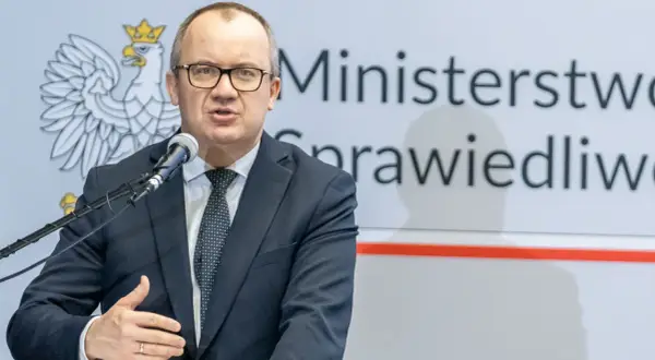 Mężczyzna w garniturze przemawia przy mikrofonie na tle godła Polski i napisu "Ministerstwo Sprawiedliwości". Może mówić o problemach z mieszkaniem komunalnym w Warszawie.