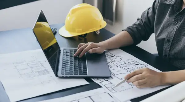 Architekt analizuje plany, obliczając powierzchnię użytkową budynku na laptopie. Obok leży żółty kask.