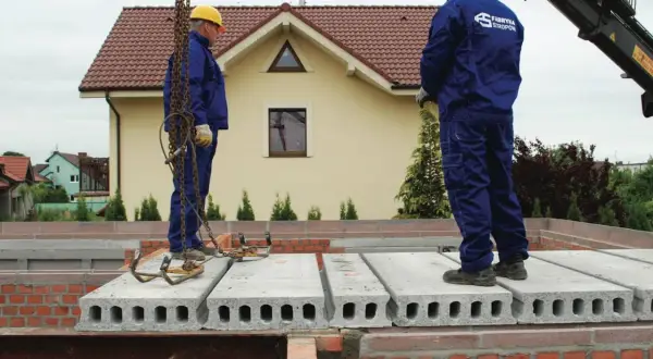 Dwóch robotników w żółtych kaskach i niebieskich kombinezonach montuje strop filigran na budowie domu.