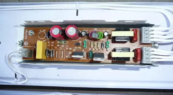 Elektroniczny statecznik z kondensatorami, cewkami i innymi elementami na płytce drukowanej, podłączony białymi przewodami.