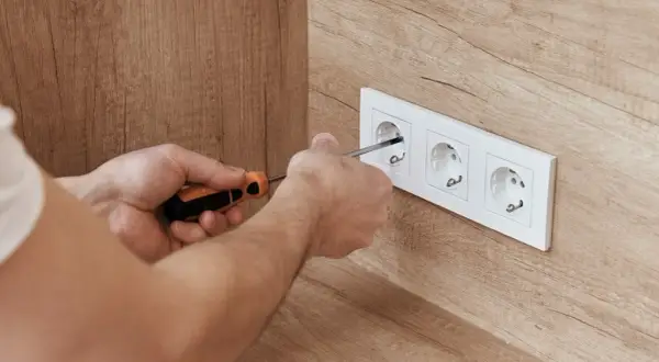 Ręce majsterkowicza dokręcają śrubki w białej ramce z trzema gniazdkami, zapewniając bezpieczne napięcie w gniazdku.