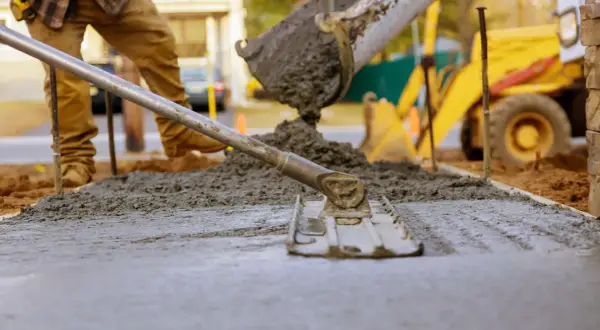 Pracownik wylewa beton na chudziaka, używając łaty do wyrównania powierzchni. W tle widać koparkę.