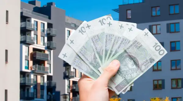 Dłoń trzyma polskie banknoty 100 zł na tle nowoczesnych bloków mieszkalnych. Szybka sprzedaż mieszkania przed upływem 5 lat.