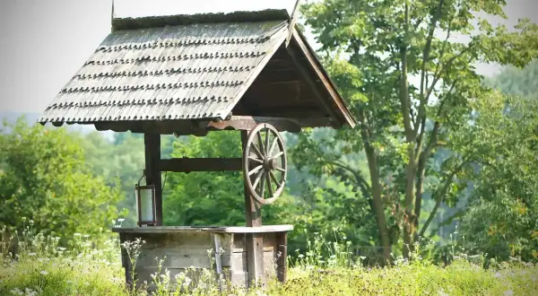 Drewniana studnia głębinowa z kołem i latarnią, otoczona polnymi kwiatami. Zastanawiasz się nad studnia głębinowa koszt?
