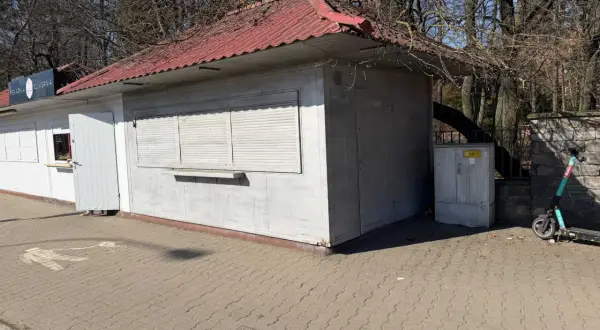 Mały lokal użytkowy z czerwoną, falistą dachem i białymi okiennicami. Obok stoi hulajnoga elektryczna.