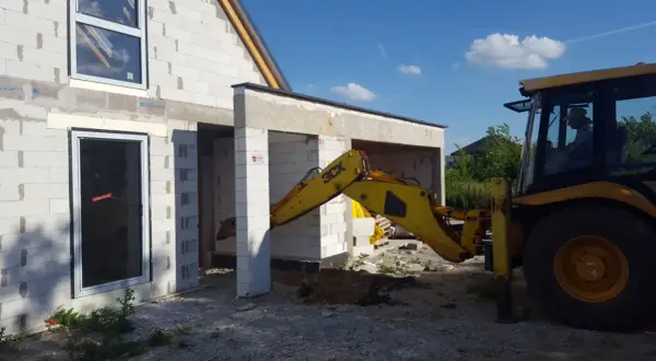 Żółta koparko-ładowarka pracuje przy budowie domu z bloczków. Widać okna i drzwi. Trwa wlz.