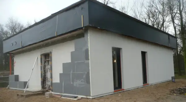 Budowa domu ze styropianu w toku. Ściany z bloczków, częściowo otynkowane, dach przykryty czarną folią.