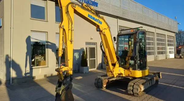 Żółta koparka Komatsu przed budynkiem. Zastanawiasz się, ile kosztuje godzina pracy koparki? Sprawdź ofertę wypożyczalni!